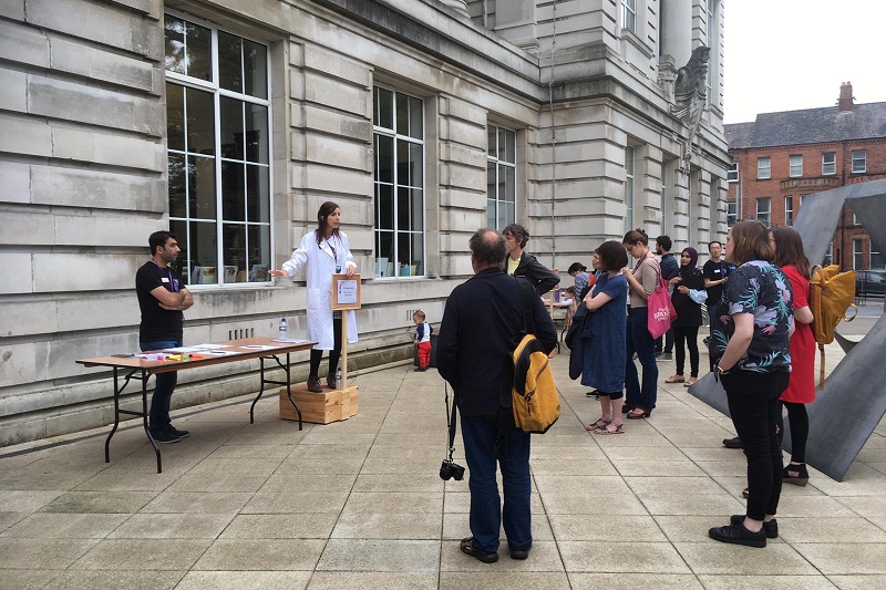 female researcher in white lab coat standing on a soapbox outside the Ulster Museum presenting to a gathered audience of spectators